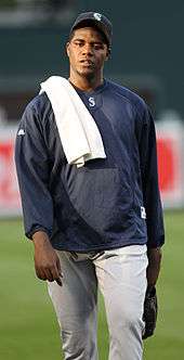 A man in a blue shirt and grey pants with a white towel over his right shoulder walks on a baseball field.
