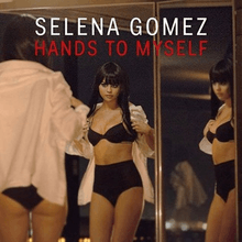 An image of Selena Gomez in a black lingerie two-piece trying on a white shirt while looking at her reflection through a glass door.