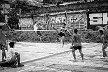 Several men playing a ball game; two of them jumping mid-air on either side of a net about eye-level high, appearing to be kicking the ball over the net; graffiti on a wall in the background