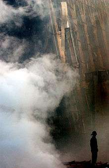 Photograph of smoke rising in front of upright steel members of a shattered building. A firefighter is visible as a dark silhouette standing still amid the smoke.