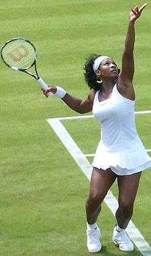 An African American female tennis player standing on a grass court wearing a white dress holding a tennis racket in her left hand waiting for the ball to drop so she can complete her serve