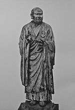 Seshin. Front view of a lifelike statue. Both arms are bend with the palm of the left hand turned upward and the right hand pointing up. Black and white photograph.
