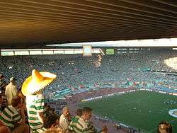 A top-stand perspective of one end of a stadium packed with people wearing white and green shirts as a football match takes place in the pitch. In the foreground, one of these people is wearing a Mexican hat.