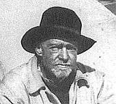  Head and shoulders of an unshaven man in dark, wide-brimmed hat, weatherbeaten face looking directly at the camera