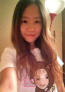 A young Asian woman with long hair wearing a white T-shirt with a stylized picture of a woman's face visible. Her lower right arm seems to extend to the camera, and behind her a wall lamp is visible.
