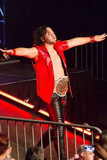 Shinsuke Nakamura, a black-haired Japanese man wearing a red vest, black pants and the white-and-gold IWGP Intercontinental Championship, struts down the entrance ramp