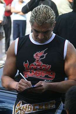 A man with bleached blond hair and wearing street clothes, signs an autograph.