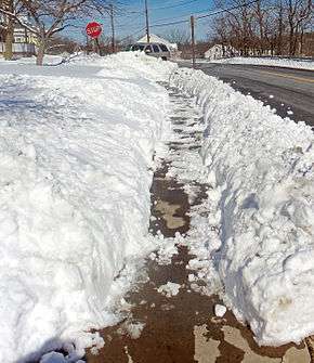 A sidewalk, covered with thin snow in many areas, with deep snow on the sides gouged irregularly. A stop sign is visible in the rear