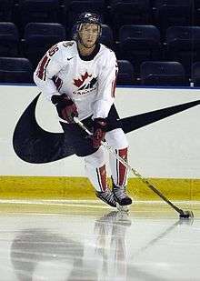 A hockey player in white Canada uniform skates across the ice, guiding the puck with his stick.