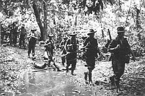 Ten men walking in single file though the jungle wearing slouch hats and carrying rifles.