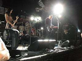 The Smashing Pumpkins perform on a brightly lit stage with a large metal flower hanging over them. From left to right: Nicole Fiorentino—a brunette Caucasian woman wearing a dress—looks at her black bass guitar, Mike Byrne plays a silver drum kit obscured behind his drums, Billy Corgan—a bald, middle-aged Caucasian man wearing a sriped t-shirt and dark pants—plays guitar, and Jeff Schroeder—a Korean male in his 30s with brunette hair—looks at his guitar while playing.