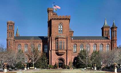 A brick building, reminiscent of a castle, slightly obscured by bare trees. There is a large central tower with the entrance at its base as well as smaller tower at each corner of the building, each with a varying design.