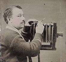 Dark-haired man with small moustache; operating large bellows camera