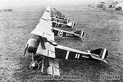A side-on view of a line of several triplane aircraft. All are painted in dark colours, with white numbers on the side.