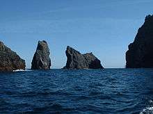 A series of dark black rocky shapes traverse from left to right in a dark blue sea under pale blue skies. The four main structures are tall and cliff-girt and set at odd angles to one another – the shapes are suggestive of a gathering of living creatures taken from a bestiary.