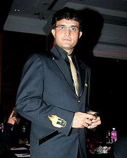 Sourav Ganguly wearing glasses stares directly at the camera. He is wearing a black suit and a gold tie. The man's right arm is facing the camera. The arm has a small picture of a helmet engulfed with fire.