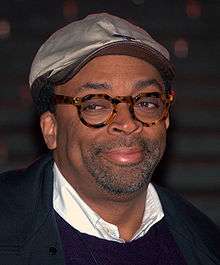 A headshot of a middle-aged African American man. Wearing round glasses and a silver cap, the man sports dark stubble.