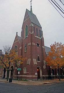 A brick church with ornate stone trim, seen from the opposite corner of an intersection. In the center is a tall steeple with a steeply gabled roof and cross on top; the lower sanctuary is to the side. On the sidewalk on either side is a tree with yellow leaves.