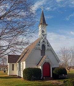 A white church with vertical siding, decorative woodwork on the roofline of its steeply gabled roof, and a red door in front, topped with an open belfry.