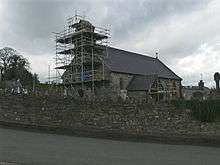 A small stone church with a slate roof see from the southwest. Scaffolding surrounds the west end and bellcote, and the porch