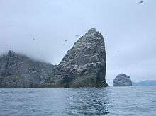 A large, triangular rock rises from the misty waters, with more islands behind and northern gannets flying around it.