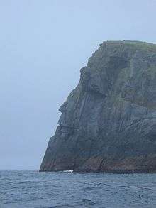 A tall grey rocky cliff towers over dark waters. The edge of the cliff is silhouetted against a leaden sky and topped with grass, creating a shape resembling a man's face.