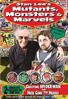A photograph of two men, both of whom are wearing glasses, looking at the viewer, and standing on a yellow-and-black-checkered floor next to a wall of comic books