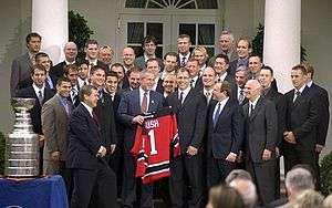 A group of young to middle-aged men stand around an older man holding a red ice hockey jersey bearing the word "BUSH" and the number "1"