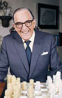 An elderly man sitting at a chess board wearing glasses smiling broadly at the camera