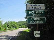 Four signs on a green, metal pole against a background of greenery. The first, which is white with a blue border and lettering, reads, "VILLAGE OF/STARLIGHT." The second and third, which are both green with a white border and lettering, read, "BUCKINGHAM/TOWNSHIP," and "PERMITS/REQUIRED," respectively. The fourth, which is white with no border and black lettering, reads, "SR 370/220."