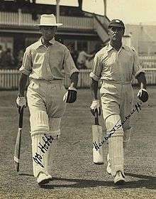 Two cricketers walking onto the field holding bats