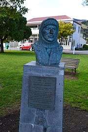 A bronze bust of a man wearing a balaclava, the bust on a plinth in a park area with a backdrop of a park bench and buildings in the far distance