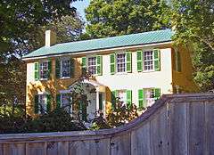 A yellow house with green shutters and roof with a wooden fence in front