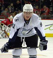 Hockey player in white and blue Tampa Bay uniform. He crouches, holding his stick on his knees.