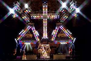 A color photograph of the band Stryper on stage under a large cross of lights