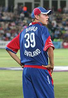An English cricketer facing away and to the right, he is wearing the red, white and blue uniform of the England cricket team and he has his hands on his hips. The word BROAD appears above the number 39 and above the word ENGLAND on the back of his shirt.