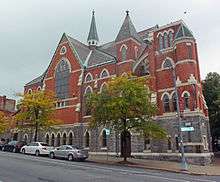 A large ornate brick church with stoned pointed-arch windows and foundation, seen from across the street