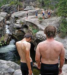 Two shirtless men in dark shorts stand with their backs to the camera. Behind them is a large area with rocky cliffs and deep water. Across it several other people similarly attired are standing