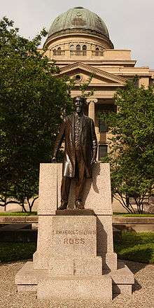A large domed building overlooks a full-length statue of balding white male with a mustache and long goatee and wearing a knee-length coat. The pedestal is engraved "Lawrence Sullivan Ross".