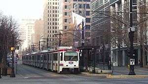 A white and red light rail train is stopped at an island platform station in the median of a street. The overhead lines powering the train are visible above the it. A row of mid- and high-rise buildings provides the background.