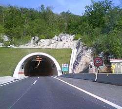  A tunnel portal with variable traffic signs indicating traffic flow direction and speed limit enforced are visible at the tunnel entrance and to the side of the road