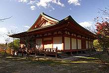 Wooden building with white walls, vermillion red beams, slightly raised floor and a hip-and-gable roof.