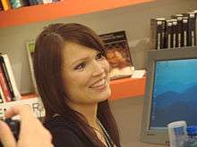A smiling Turunen is seen in front of a book shelf and a computer screen.