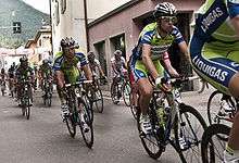 A group of road racing cyclists led down the road by several wearing matching green and blue jerseys with white trim.