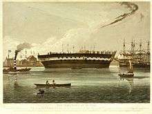 Print of the hull of a sailing ship without masts or rigging aground on mud beside a river.