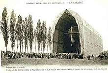 An airship inside a field hangar in centre, only the tail of the airship is visible; to the left of the hangar is a row of poplars or approximately the same height as the hangar roof. In front of the hangar a large crowd is assembled.
