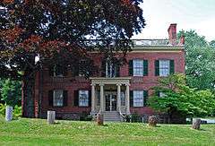 A six-bay-wide brick house with green window shutters, a balustrade on the roof and an entrance porch with round fluted Ionic columns. Along the front lawn, which rises slightly, is a line of five tree stumps with bark removed. There is a large tree on the left partially shading the house.