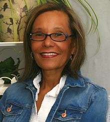 Indoor photo of a middle-aged female wearing glasses, a denim jacket and white blouse looking into the camera