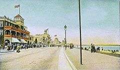An early 20th century postcard view of a beach with a road running alongside. Across the road from the beach are some buildings and a wide sidewalk full of people. There are also people standing and walking on the beach.