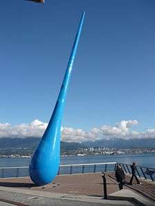 A giant raindrop, at a jaunty angle, deep blue in colour, sits on a block-paved promenade overlooking water, with mountains in the background.